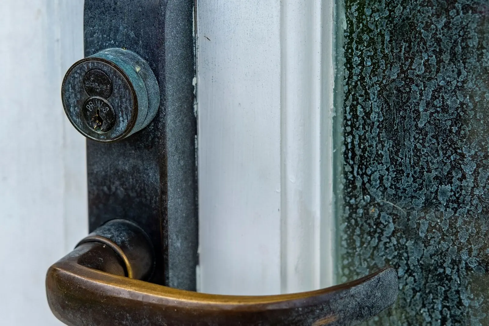 A weathered mortise lock trim on a white door frame: a round key cylinder above a curved bronze lever.