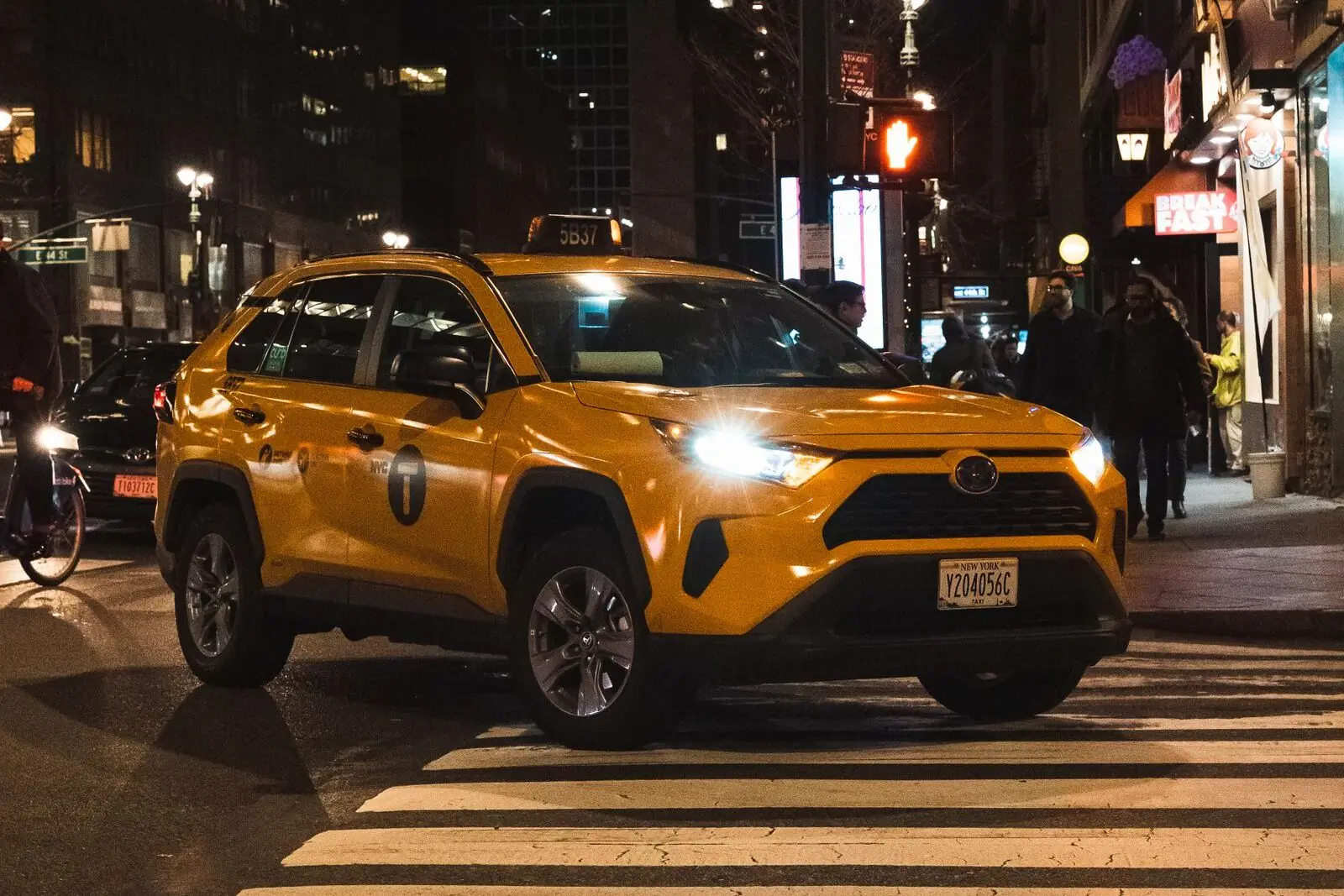 A yellow New York City taxi with its headlights on, stopped at a crosswalk on a Manhattan street at night.